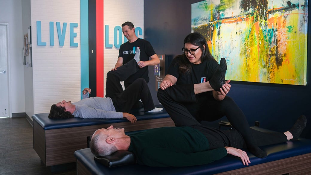 young man in 20s and older man in 50s receiving stretch therapy from trained experts at an assisted stretching studio, lying flat on backs and receiving hamstring and quad stretch for tension relief