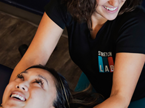 smiling woman in her 20s or 30s receiving an assisted stretch for her shoulders and neck from an expert stretch practitioner