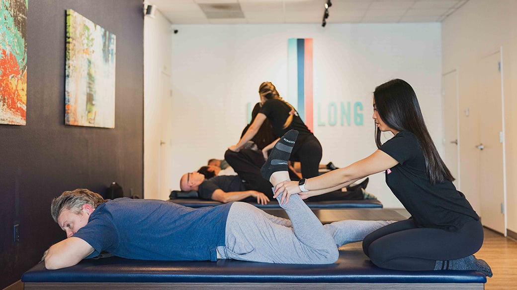 smiling fit middle aged man in athleticwear with female stretch practitioner performing piriformis stretch on glute and hamstring in bright and modern facility